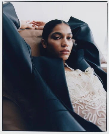 Portrait of a woman lying down wearing a lace top and black blazer. Her hair is slicked back, with a blurred background and neutral colors.