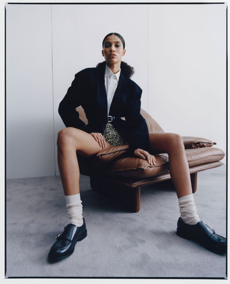 A woman sits in a lounge chair wearing a black blazer, white shirt, and patterned shorts, complemented by socks and black shoes.