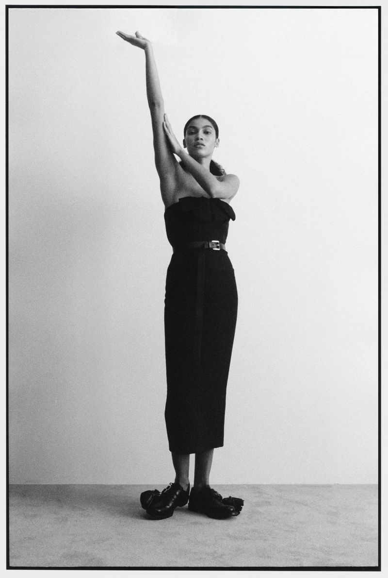 Model Raynara Negrine posing in a black dress with one arm raised against a grey seamless background