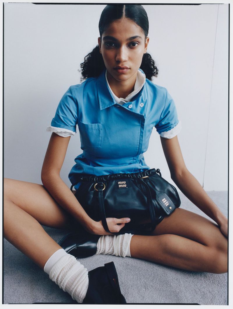 Fashion portrait of an adult female in a blue uniform-inspired dress with a black leather purse, socks, and shoes, sitting on a grey surface.