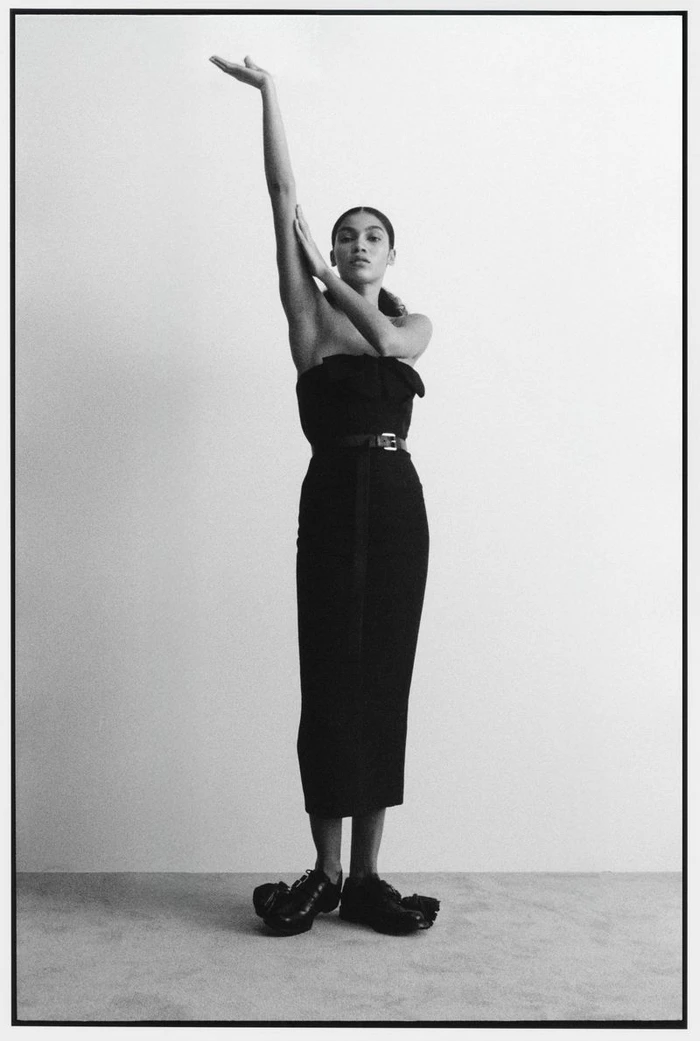 Model Raynara Negrine posing in a black dress with one arm raised against a grey seamless background