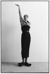 Model Raynara Negrine posing in a black dress with one arm raised against a grey seamless background