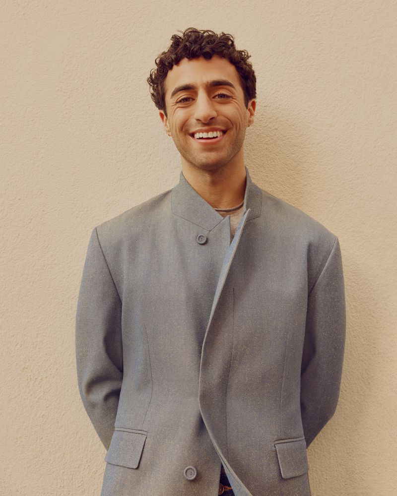 A smiling man with curly hair wearing a blue suit jacket stands against a neutral wall. The shot is a medium shot.