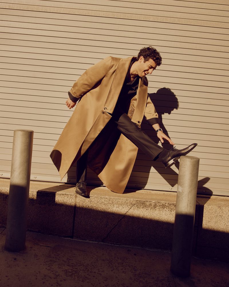 A stylish adult male, wearing a tan overcoat and black dress pants, strikes a dynamic pose on a city street with light and shadow play.