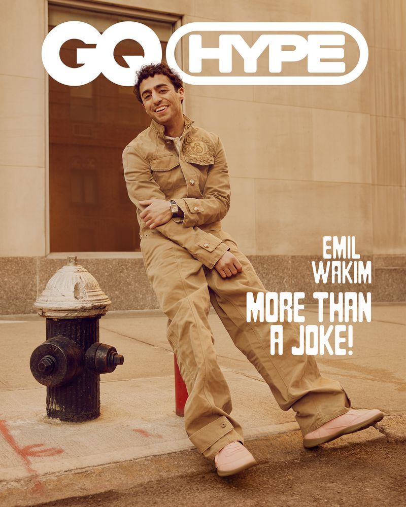Emil Wakim poses for a GQ Hype cover. He is sitting on a red fire hydrant. He is wearing a tan jacket and pants. A building is in the background.