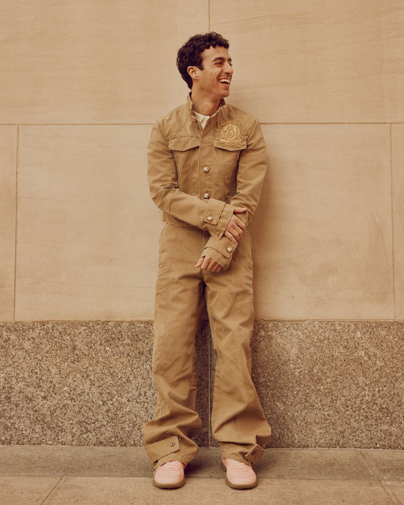 Smiling man in tan jumpsuit and pink shoes stands against a neutral urban wall on a sunny day, radiating a casual yet stylish vibe.