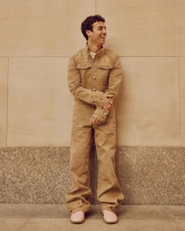 Smiling man in tan jumpsuit and pink shoes stands against a neutral urban wall on a sunny day, radiating a casual yet stylish vibe.