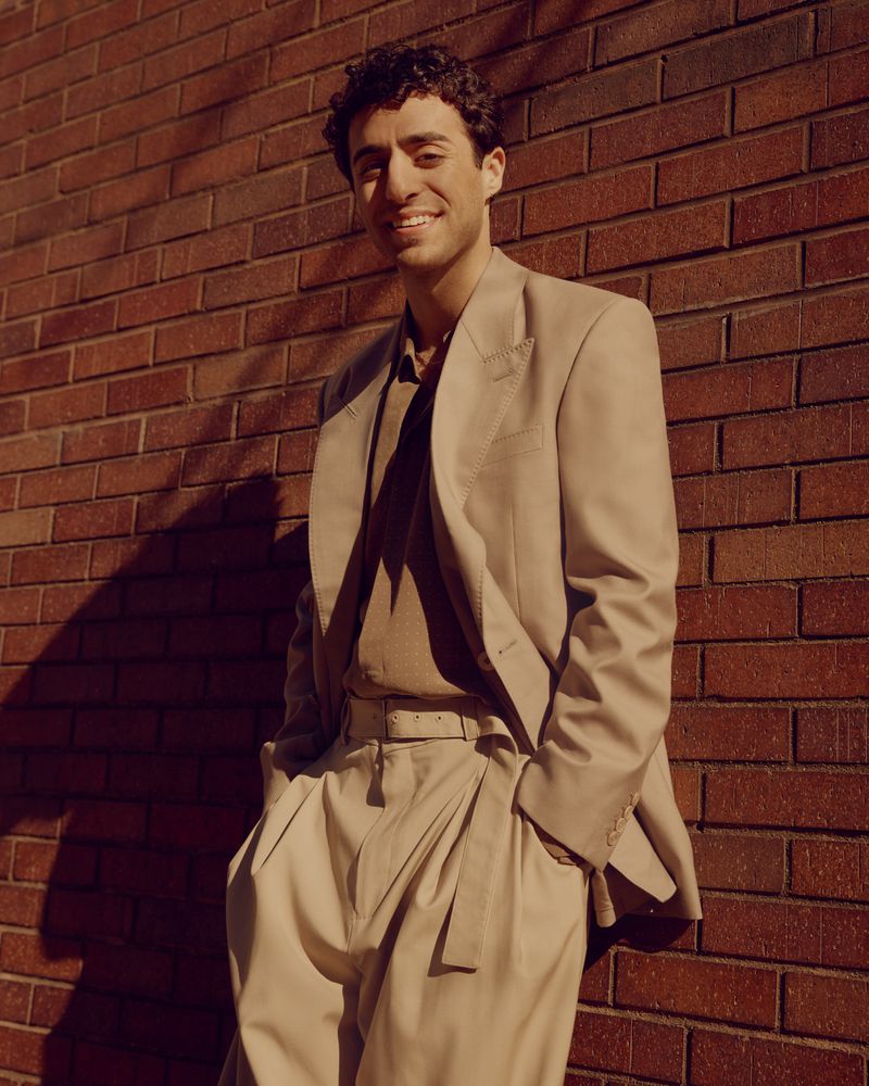 Smiling adult man in a beige suit poses against a red brick wall in the sunlight, creating long shadows in an outdoor setting.
