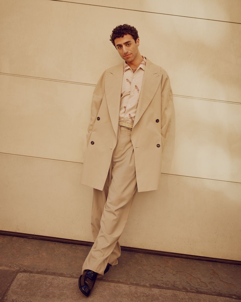 Young man in a beige double-breasted trench coat and matching trousers stands against a cream wall, striking a casual pose in an editorial fashion shot.
