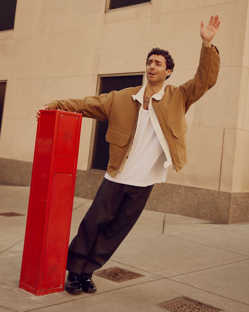 A young adult male in a tan jacket poses outdoors, leaning on a red post with one arm raised, on a city street with a neutral-colored building.
