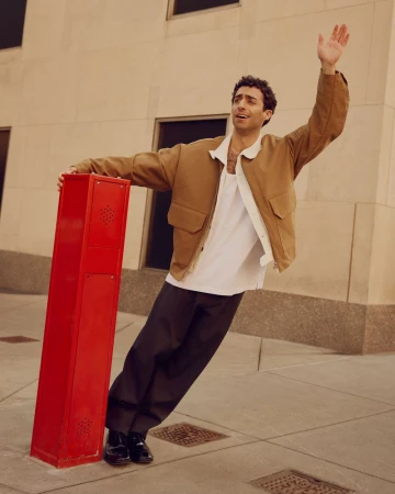 A young adult male in a tan jacket poses outdoors, leaning on a red post with one arm raised, on a city street with a neutral-colored building.