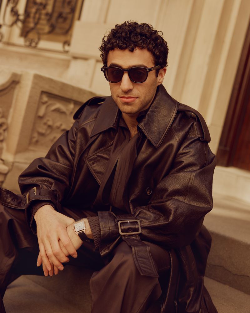 Man wearing dark sunglasses and a leather trench coat seated outdoors on stone steps for a GQ Middle East editorial portrait.