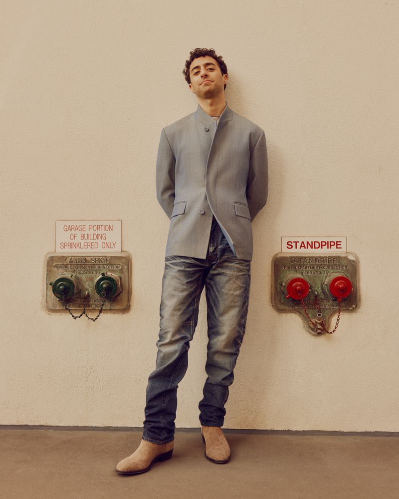Full-shot of a man standing indoors in a grey jacket and jeans, with metal fire safety equipment on the wall behind him.