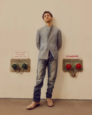 Full-shot of a man standing indoors in a grey jacket and jeans, with metal fire safety equipment on the wall behind him.