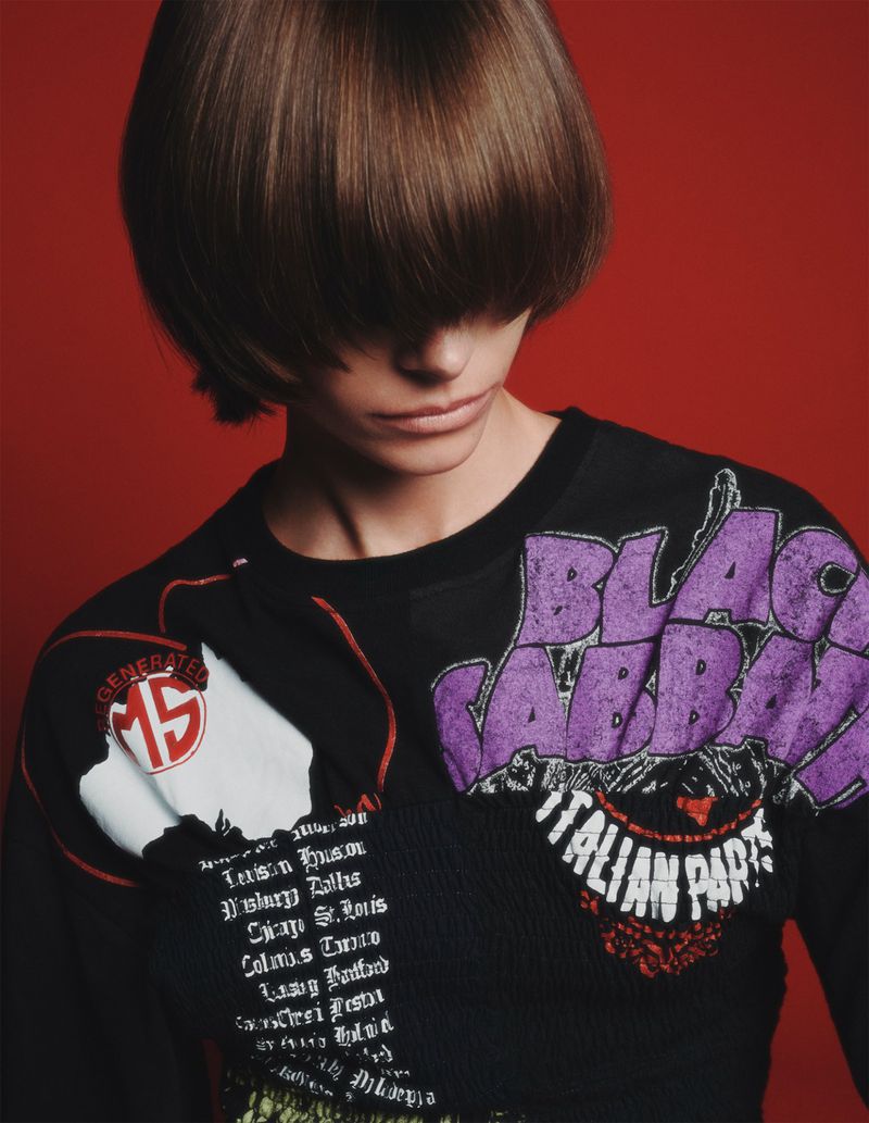 A person with brown hair poses in front of a red background, wearing a black shirt with band logos and text.