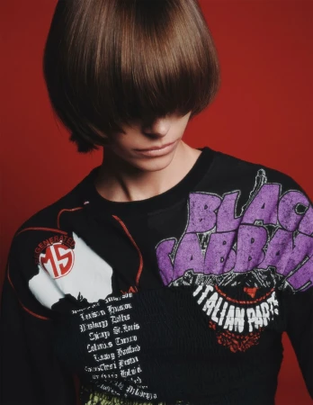 A person with brown hair poses in front of a red background, wearing a black shirt with band logos and text.