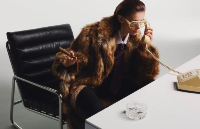 A woman in a fur coat sits at a desk, talking on a vintage phone while holding a cigar, indoor in a staged photo studio environment.