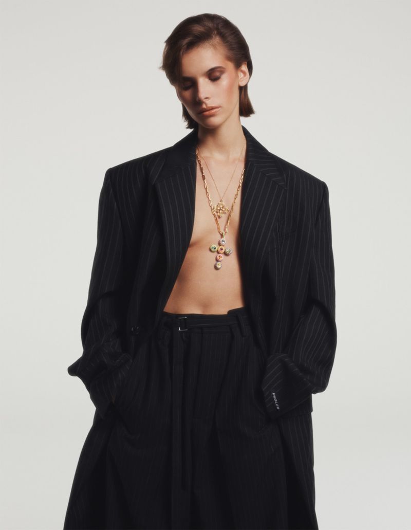Fashion portrait of a woman in an oversized pinstripe blazer and wide-leg trousers, accessorized with layered necklaces against white.