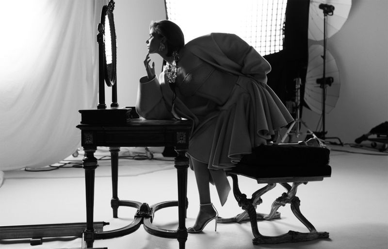Black and white photo of a woman in a coat seated at a vanity, looking at her reflection in the mirror in a photo studio.
