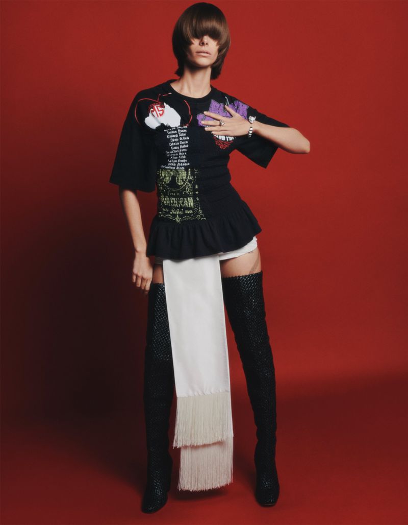 A woman with dark, short hair poses in a black printed t-shirt, white shorts, and thigh-high sparkly boots against a red backdrop.