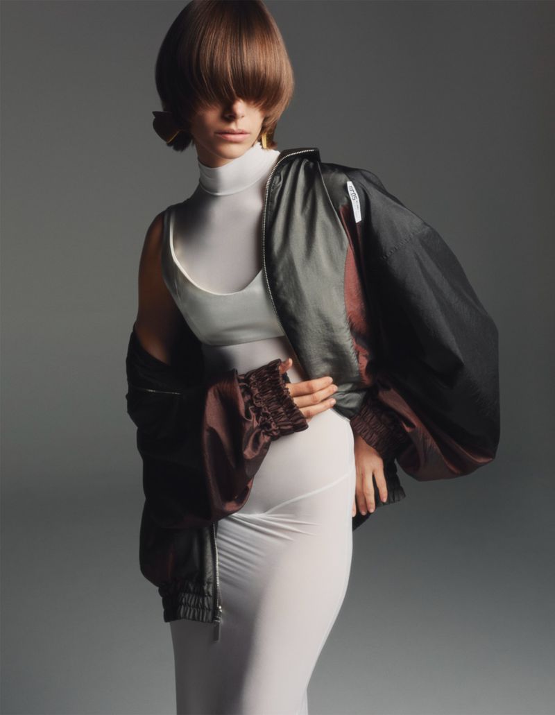 Woman in a white top and fitted skirt poses in front of a grey background, wearing a black and brown bomber jacket, studio lighting.