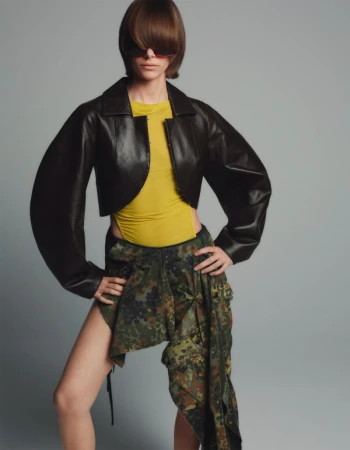 Model wearing a black leather jacket, yellow top, and camo skirt against a grey background for Sixteen Journal.