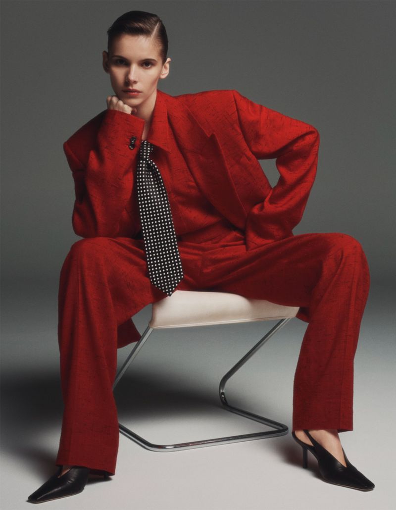 Model wearing a red suit and polka dot tie seated on a chair against a grey studio backdrop