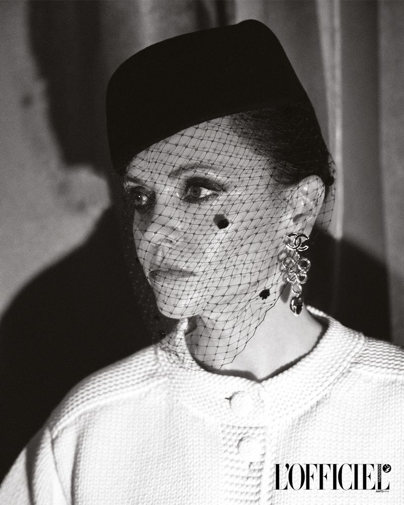 Black and white portrait of an adult woman with a hat and netting over her face, wearing a tweed jacket, and large flower earrings.