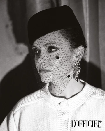 Black and white portrait of an adult woman with a hat and netting over her face, wearing a tweed jacket, and large flower earrings.