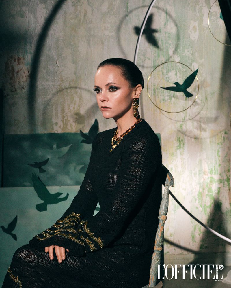 Elegant woman in dark dress and gold jewelry poses seated in a studio with bird silhouettes in the background in an eye-level medium shot.