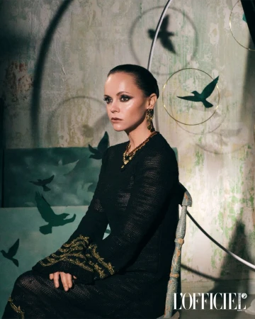 Elegant woman in dark dress and gold jewelry poses seated in a studio with bird silhouettes in the background in an eye-level medium shot.