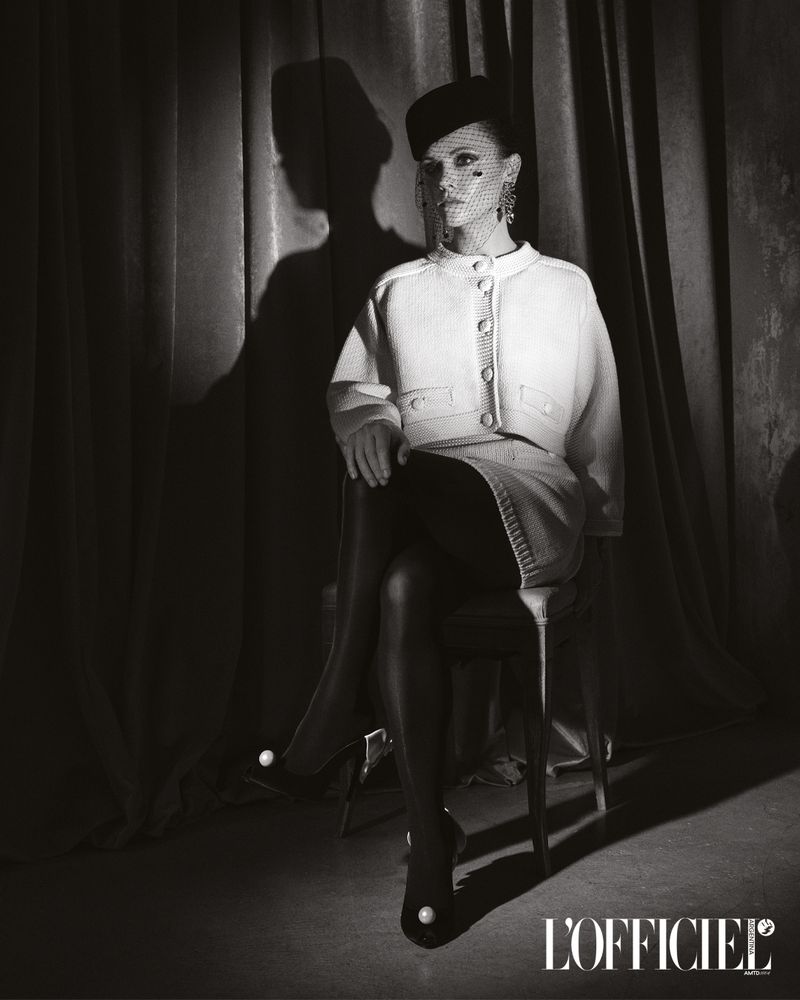 Elegant woman in a white skirt suit, fishnet veil, and pearl-embellished heels, seated against a neutral curtain backdrop in a studio setting.