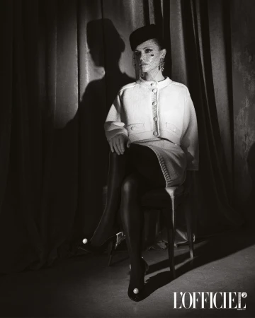 Elegant woman in a white skirt suit, fishnet veil, and pearl-embellished heels, seated against a neutral curtain backdrop in a studio setting.