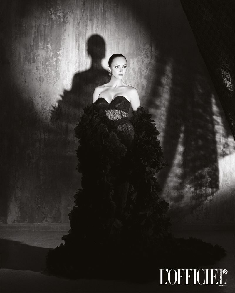Black and white portrait of Christina Ricci wearing a strapless black gown with voluminous ruffled stoles against a textured wall with shadows.