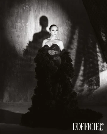Black and white portrait of Christina Ricci wearing a strapless black gown with voluminous ruffled stoles against a textured wall with shadows.