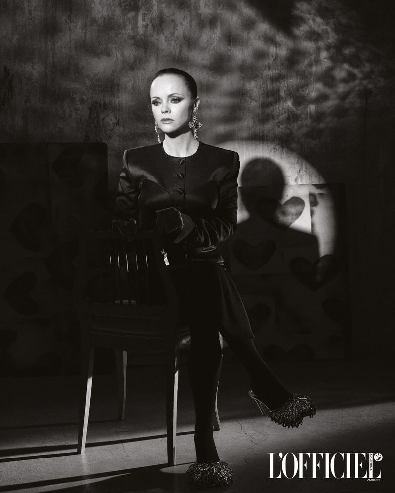Black and white portrait of Christina Ricci in a dark jacket and heels, seated against a textured wall with a spotlight.