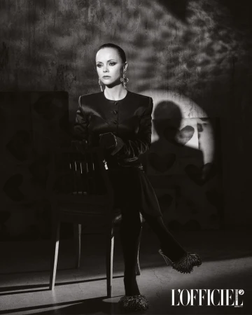 Black and white portrait of Christina Ricci in a dark jacket and heels, seated against a textured wall with a spotlight.