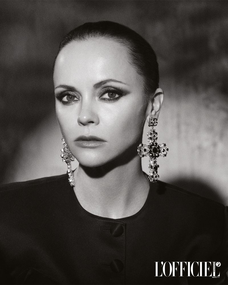 Striking black and white portrait of Christina Ricci in an elegant black top and ornate cross earrings against a textured backdrop.