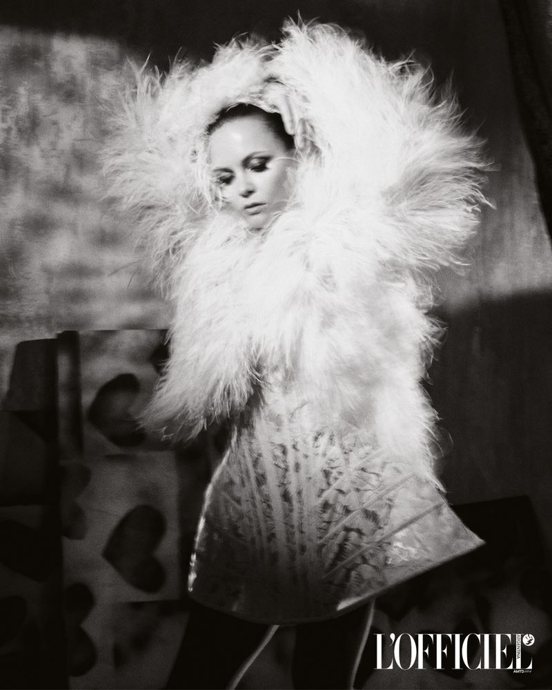 Black and white fashion shot: a female model wears a feathered coat and dress, holding her hands on her head, in a photo studio.