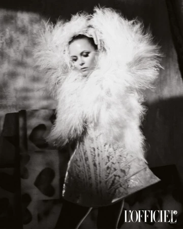 Black and white fashion shot: a female model wears a feathered coat and dress, holding her hands on her head, in a photo studio.