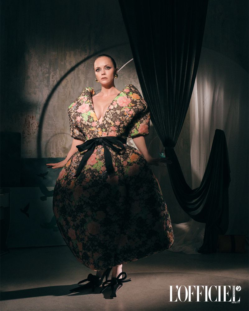 A woman poses in a floral dress with puffed sleeves and a bow at the waist, in a studio with draped fabric and L'Officiel logo.
