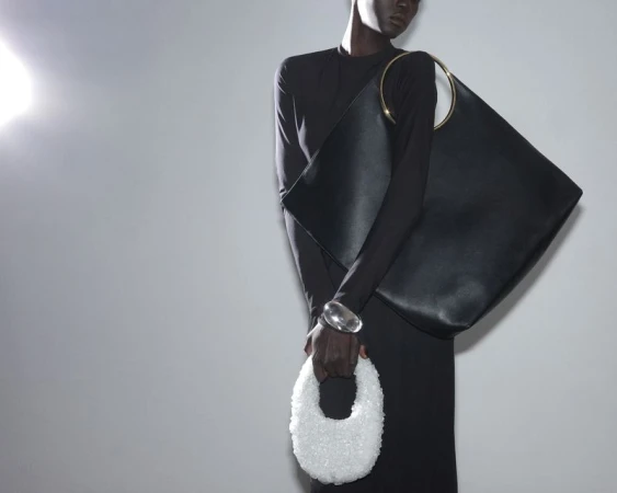 Fashion shot of a Black woman wearing a long-sleeved black dress, silver bracelet, and holding a large black purse and a beaded white purse.