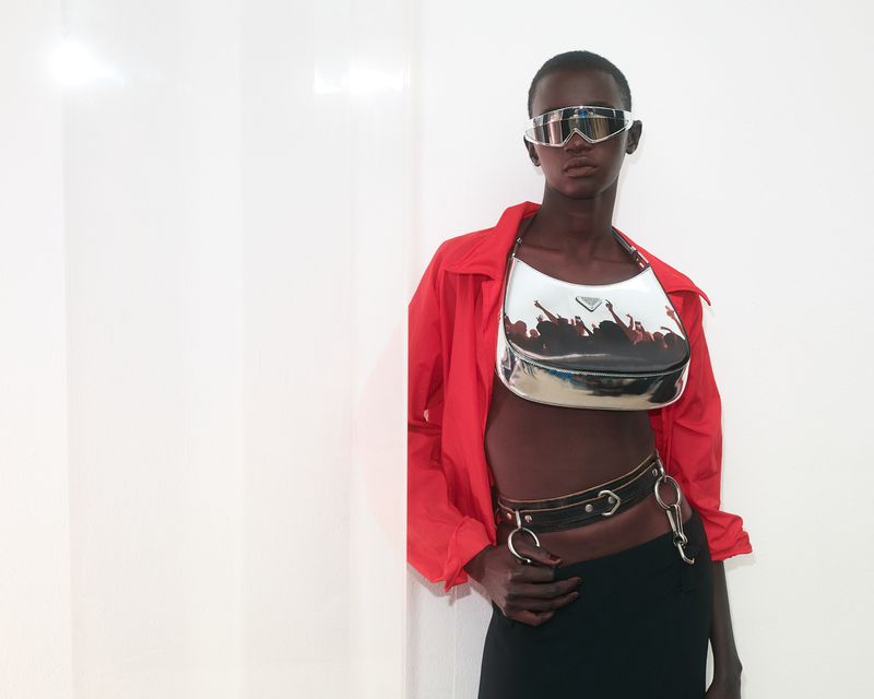 Model in a red shirt, black skirt, metallic glasses, and a silver Prada purse poses against a white wall in a fashion editorial.
