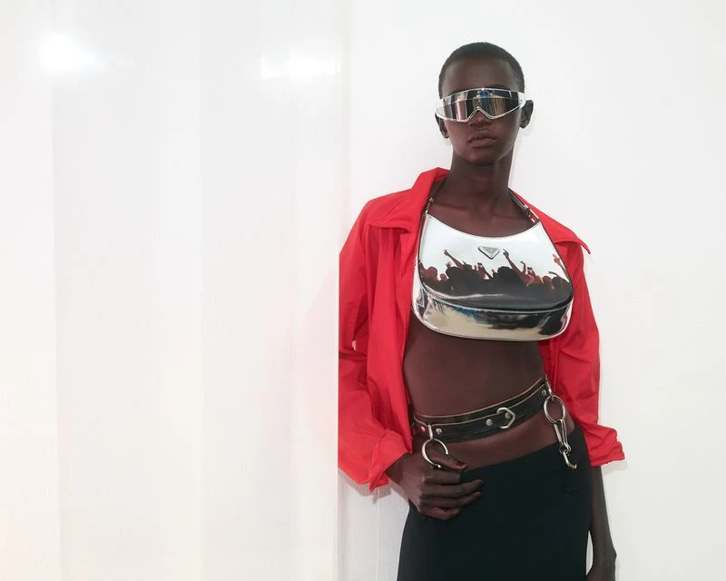 Model in a red shirt, black skirt, metallic glasses, and a silver Prada purse poses against a white wall in a fashion editorial.