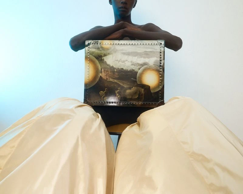 A bare-chested, dark-skinned person seated on a chair is draped with ivory fabric. Leaning against an art-covered trunk, studio lit.