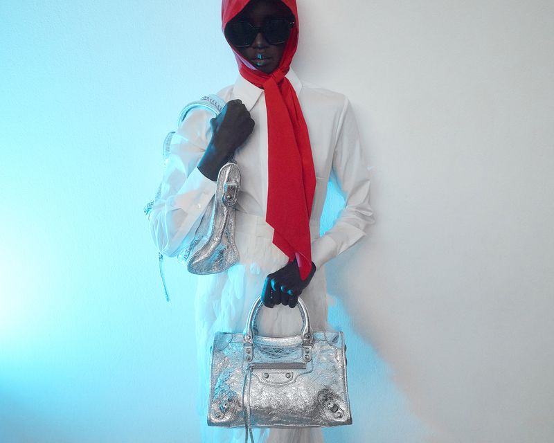 Dark-skinned model in white shirt and trousers, red headscarf, and sunglasses, holding a silver handbag in a fashion photography studio.