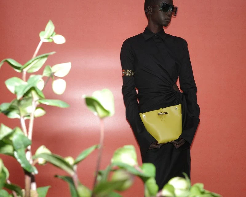 An adult fashion model poses with a chartreuse handbag in a studio setting, accented by leafy foliage and wearing an all-black outfit.