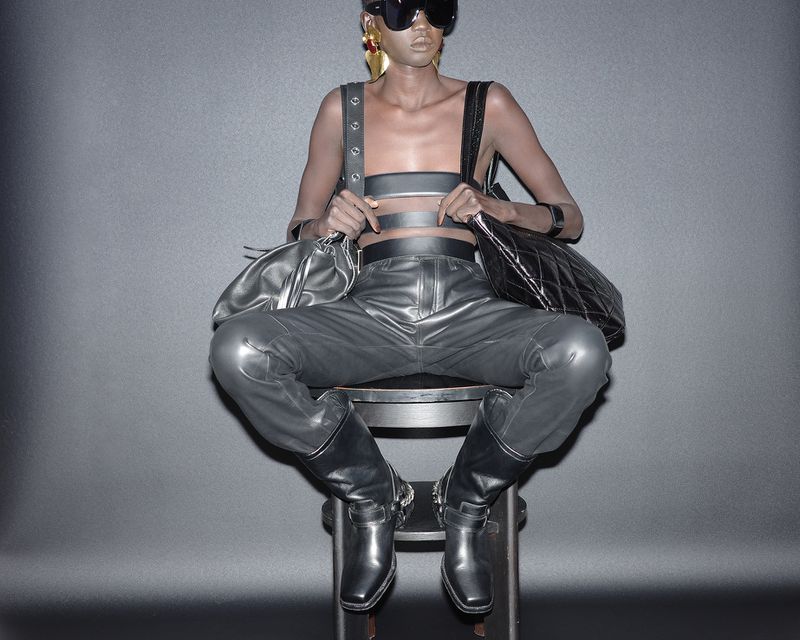A model in sunglasses and avant-garde fashion sits on a stool against a gray backdrop with a leather top, pants, boots, and accessories.
