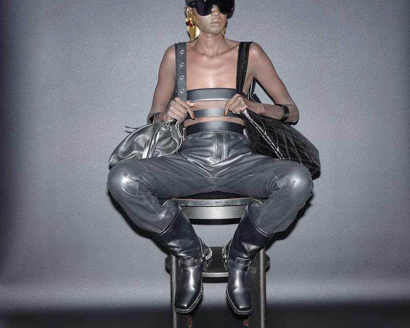 A model in sunglasses and avant-garde fashion sits on a stool against a gray backdrop with a leather top, pants, boots, and accessories.