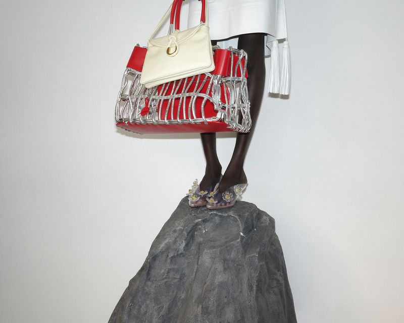 Model standing on a grey rock holding red and silver structured designer handbags against a plain white wall